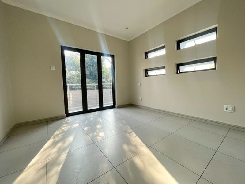 A bright, empty bedroom featuring large aluminium sliding doors leading to a balcony and modern horizontal clerestory windows with neutral tiled flooring.