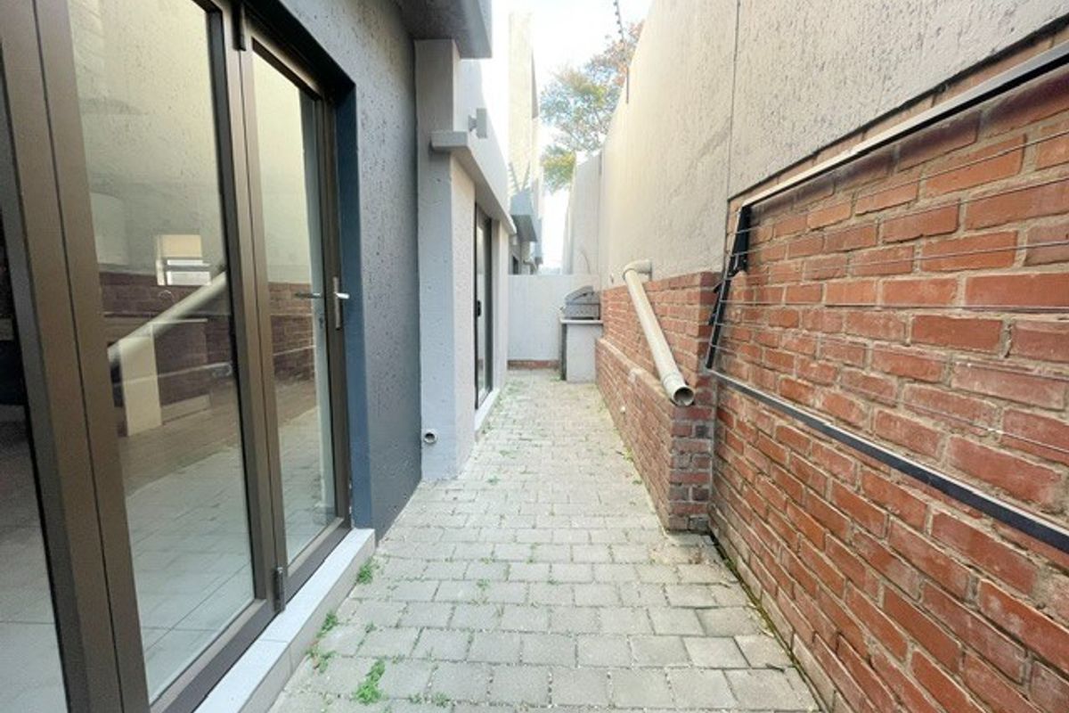 A narrow rear exterior walkway featuring paved flooring, a brick boundary wall, and access to the interior via sliding glass doors.