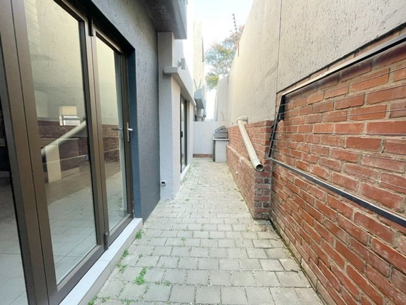 A narrow rear exterior walkway featuring paved flooring, a brick boundary wall, and access to the interior via sliding glass doors.