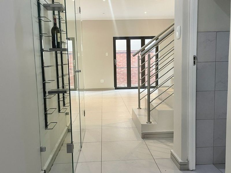 A bright, modern hallway featuring tiled flooring, a sleek stainless steel staircase railing, and a built-in glass wine display wall leading to bi-fold doors.