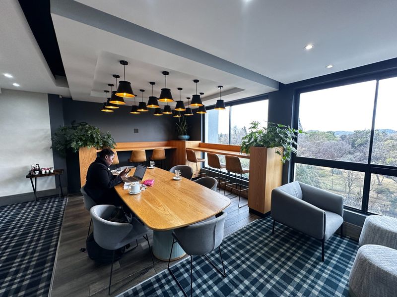 Modern open-plan communal workspace featuring a large timber table, built-in bar seating, recessed ceiling lighting, and expansive windows with natural views.