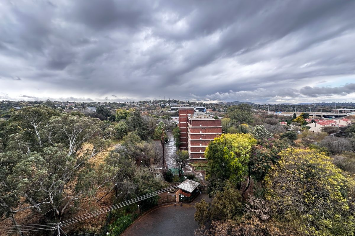 An elevated aerial view of the apartment complex grounds, featuring mature trees, a security gatehouse, and surrounding suburban landscape under a cloudy sky.