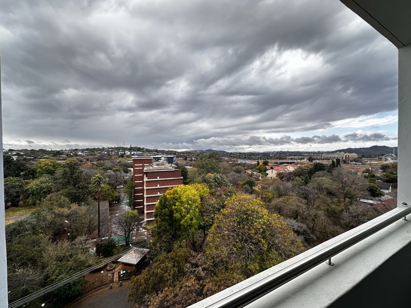 Elevated urban view overlooking surrounding neighborhood trees and buildings, captured from a balcony with a metal railing.