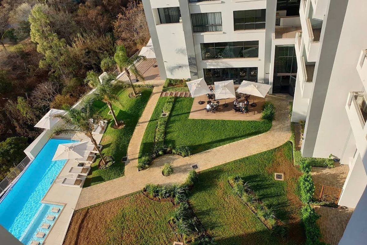 Aerial view of the apartment complex featuring a swimming pool, landscaped gardens, paved walkways, and modern building architecture.
