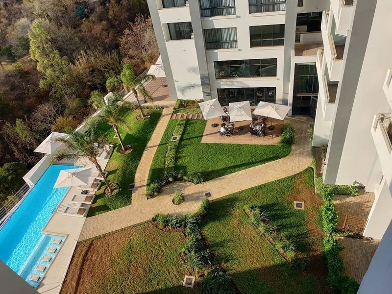 Aerial view of the apartment complex featuring a swimming pool, landscaped gardens, paved walkways, and modern building architecture.