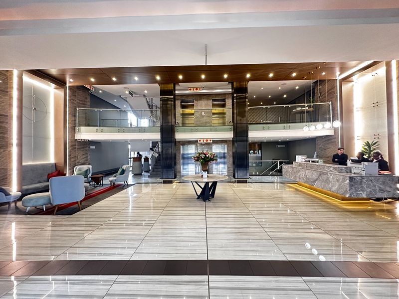 A spacious, modern apartment building lobby featuring polished marble floors, a stone reception desk, and glass-enclosed elevator access.