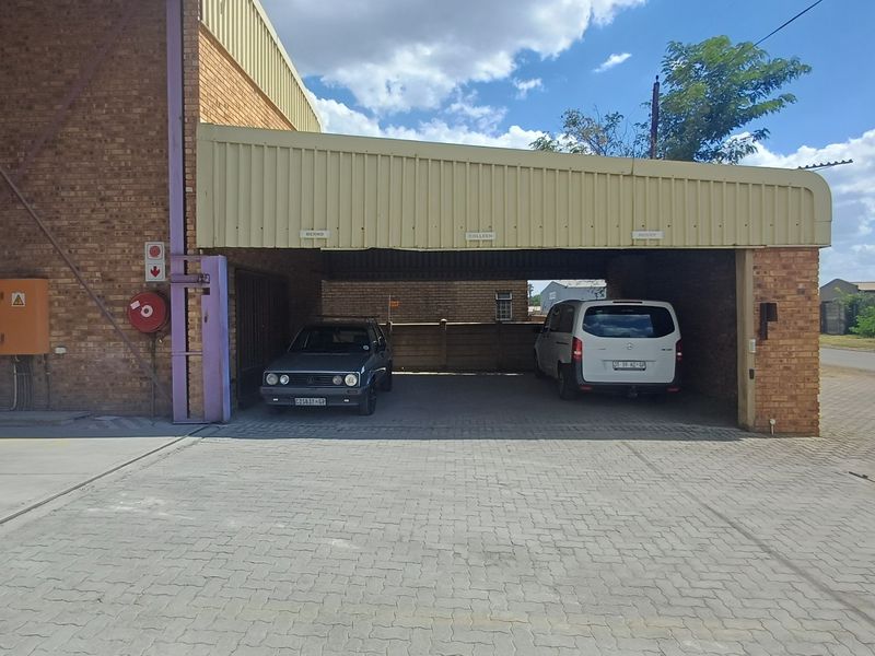 Industrial property exterior featuring a brick facade, covered parking area with metal canopy, and a paved driveway.