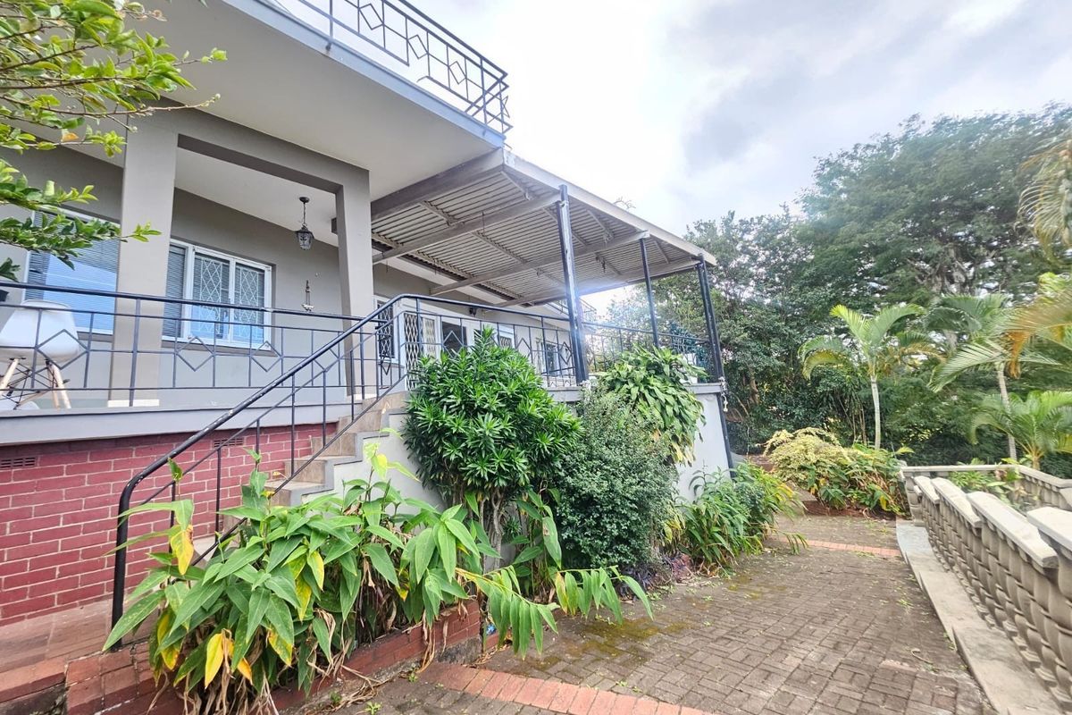 Rear exterior view featuring a covered patio, brick paved walkway, and metal balustrades leading to an upper balcony area.