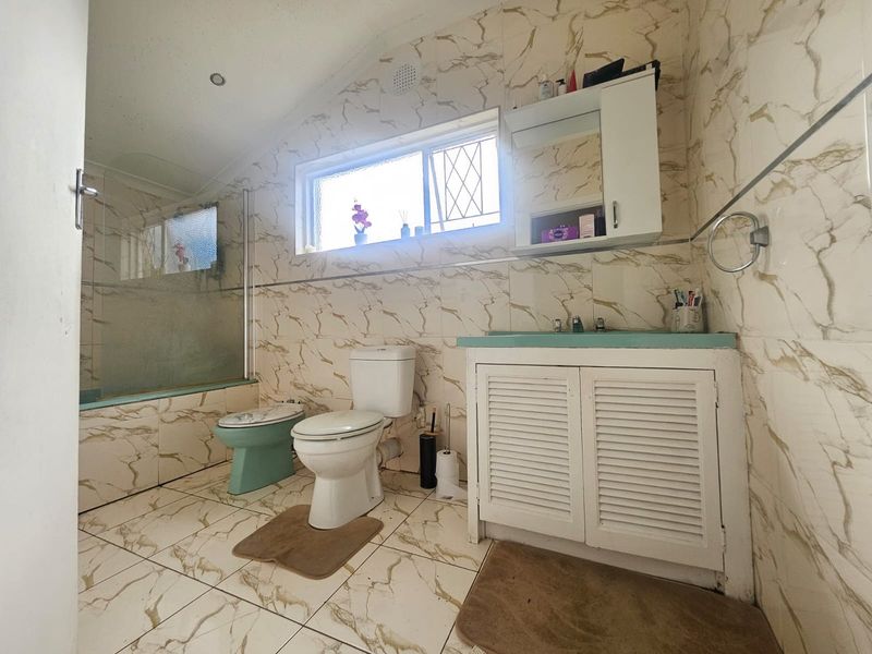 A dated bathroom featuring marble-look wall and floor tiles, a built-in vanity, toilet, and a bathtub with a glass shower screen.