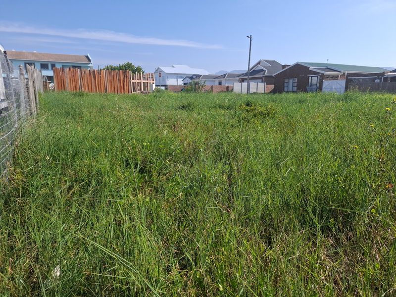 A vacant residential plot featuring overgrown grass, enclosed by wire mesh and wooden boundary fencing in a developed neighborhood.