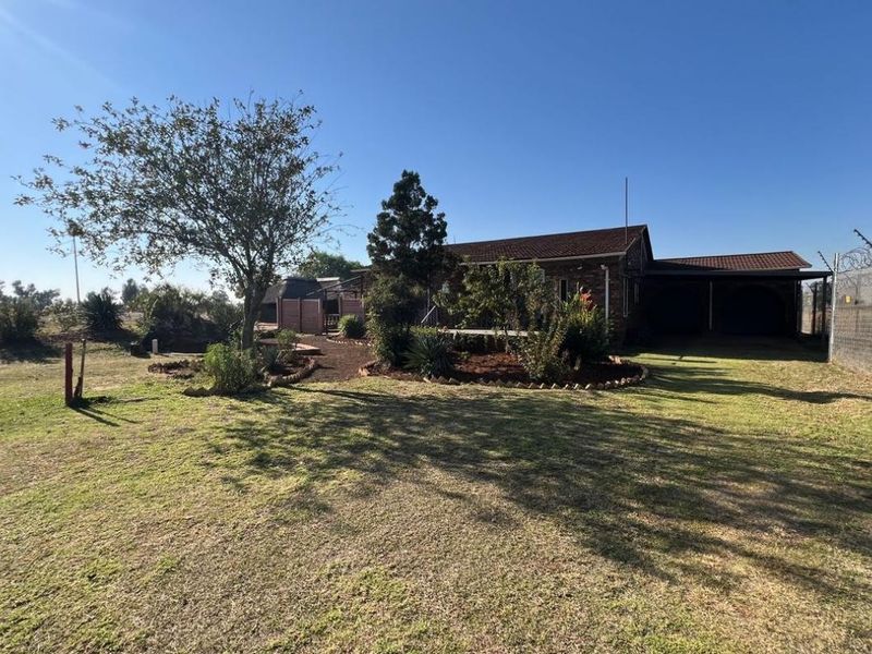 Rear view of the industrial property featuring a brick facade, tiled roof, attached carport, and established garden area with electric perimeter fencing.