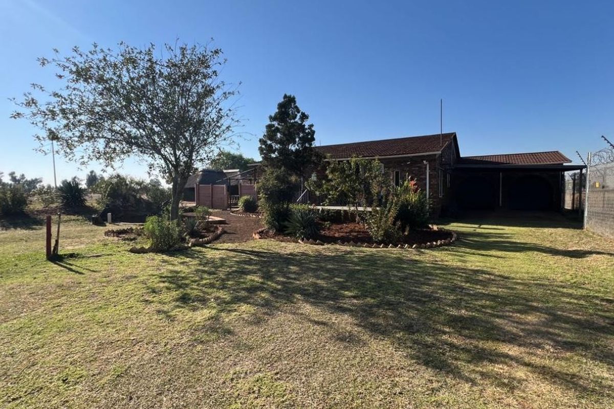 Rear view of the industrial property featuring a brick facade, tiled roof, attached carport, and established garden area with electric perimeter fencing.