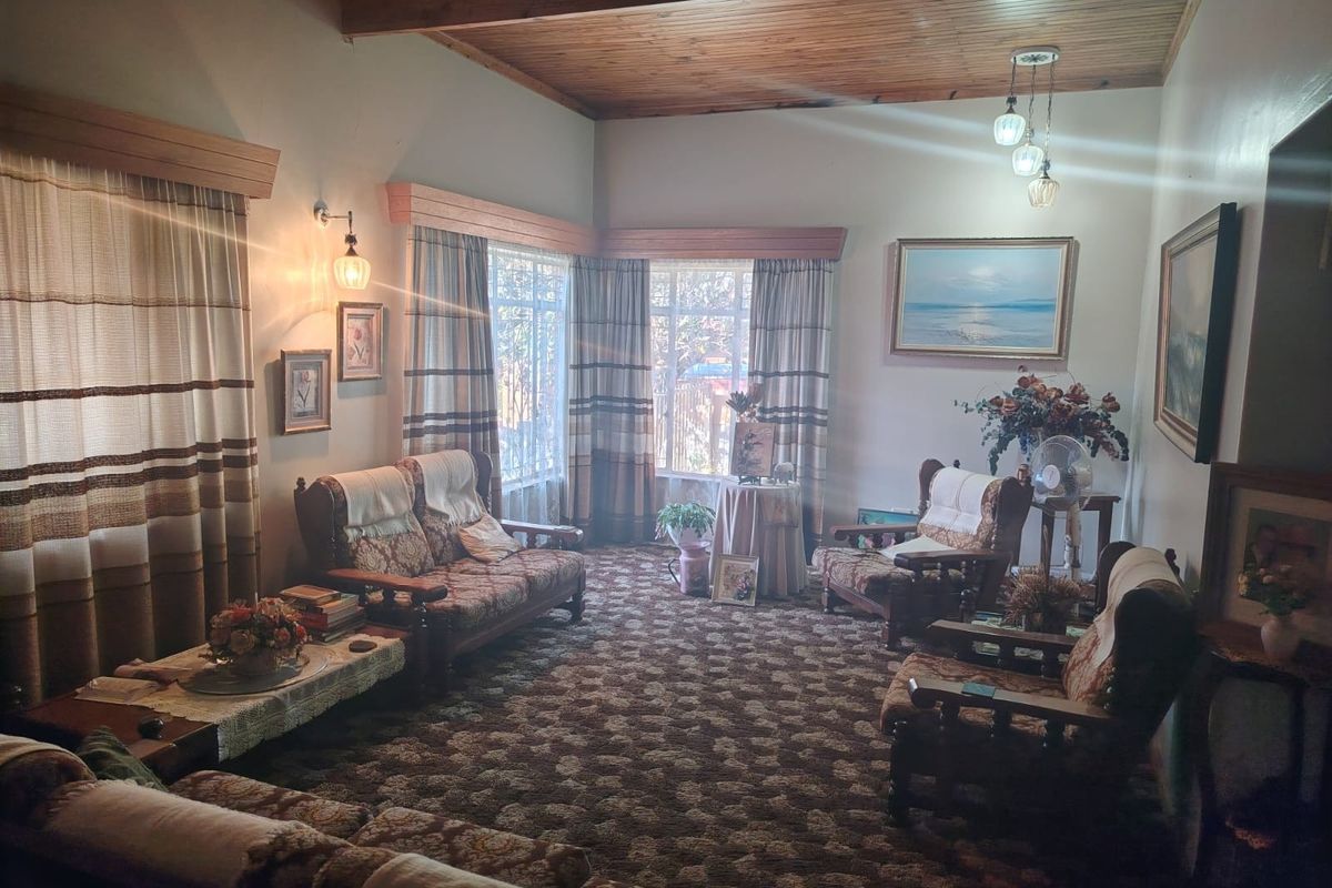 A traditional living room featuring a timber-clad ceiling with exposed beams, wall-mounted lighting, and large windows with pelmets.