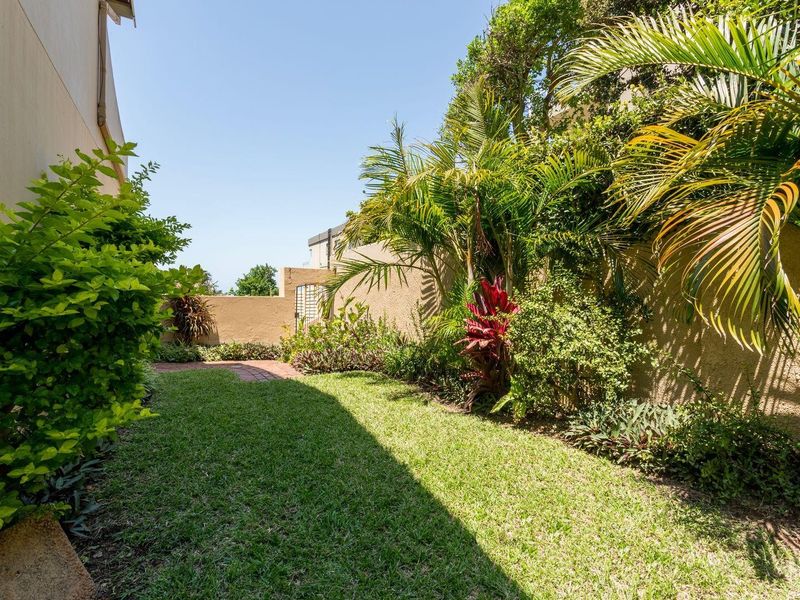 A private rear garden featuring a well-maintained lawn, established tropical landscaping, and a brick-paved pathway enclosed by boundary walls.