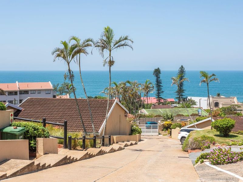 A view looking down a concrete driveway towards a security gate, featuring surrounding residential properties and a clear ocean view in the background.