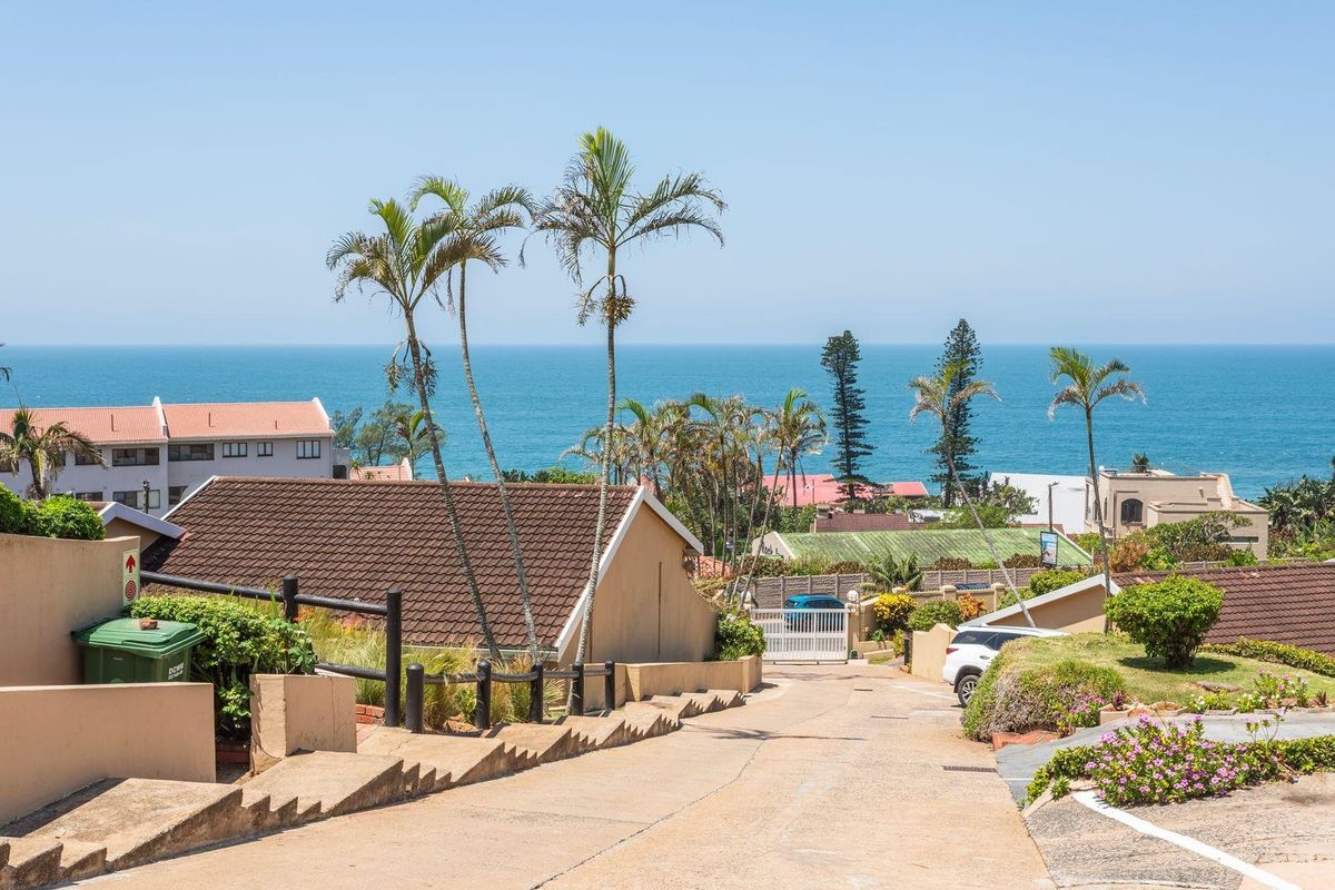 A view looking down a concrete driveway towards a security gate, featuring surrounding residential properties and a clear ocean view in the background.