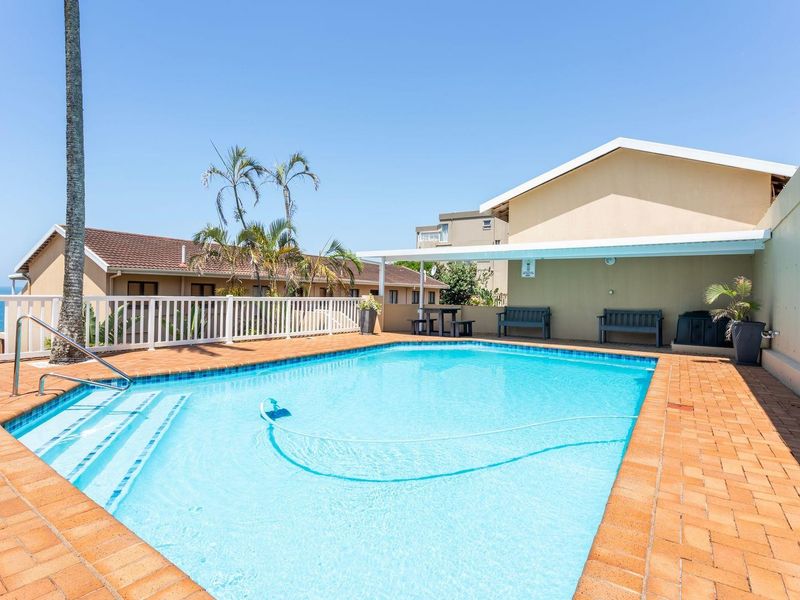 A well-maintained outdoor swimming pool area featuring a paved deck, covered patio space, and white perimeter fencing with clear views.