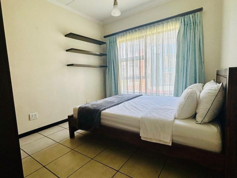 A functional bedroom featuring tiled flooring, floating wall shelves, and a window with curtain treatments, providing a neutral space for personalization.