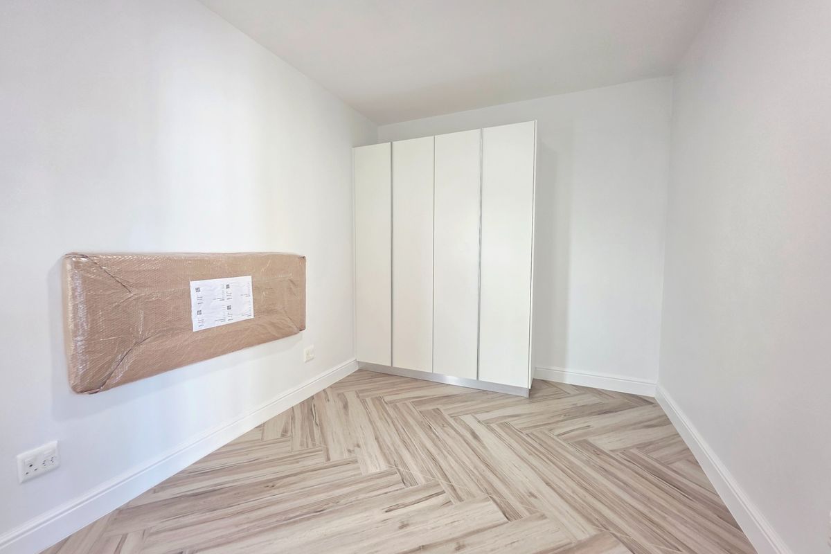 A bright, empty bedroom featuring modern herringbone wood-look flooring and a clean, white built-in wardrobe.