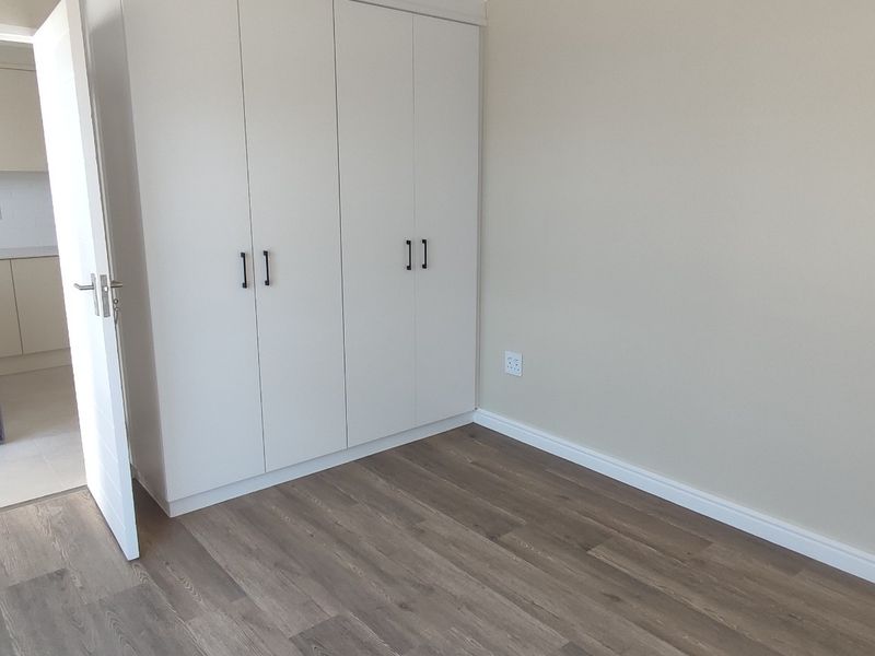 A clean, modern bedroom featuring neutral walls, durable laminate flooring, and built-in white cupboards, offering a bright and versatile living space.
