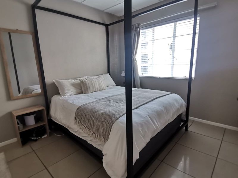 A functional bedroom featuring neutral wall tones, tiled flooring, and a window with fitted blinds, providing a simple and clean living space.