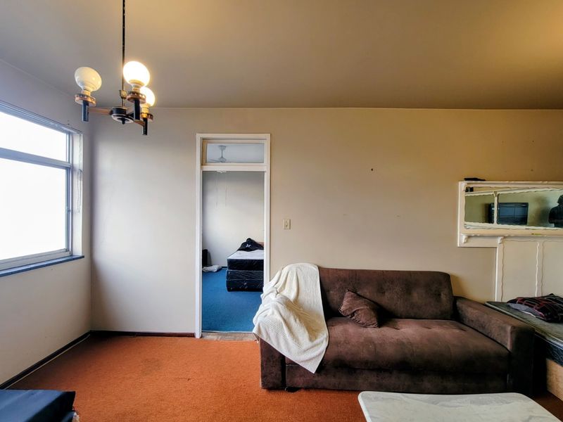 A living room featuring a large window, a ceiling light fixture, and a doorway leading to an adjacent bedroom with blue carpeting.