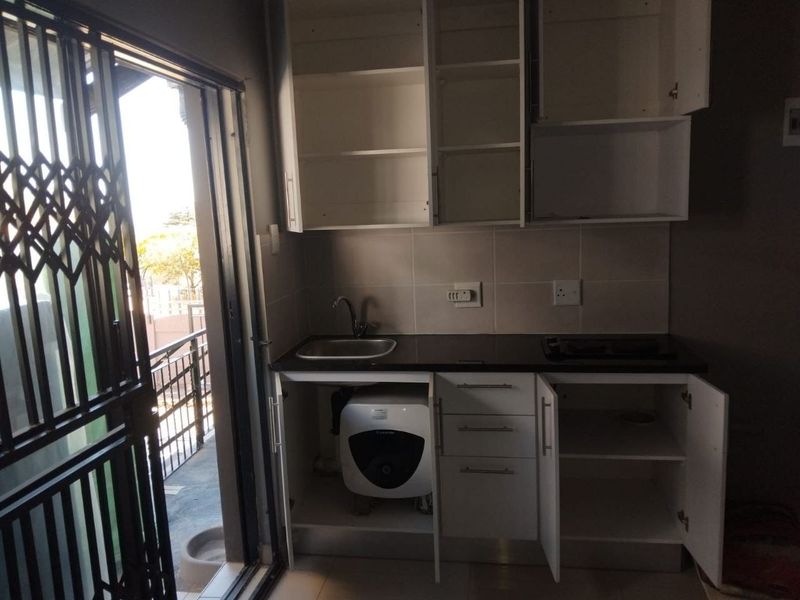 A compact kitchen area featuring built-in cabinetry, a sink, and a tiled backsplash, with direct access to an outdoor balcony through a security gate.