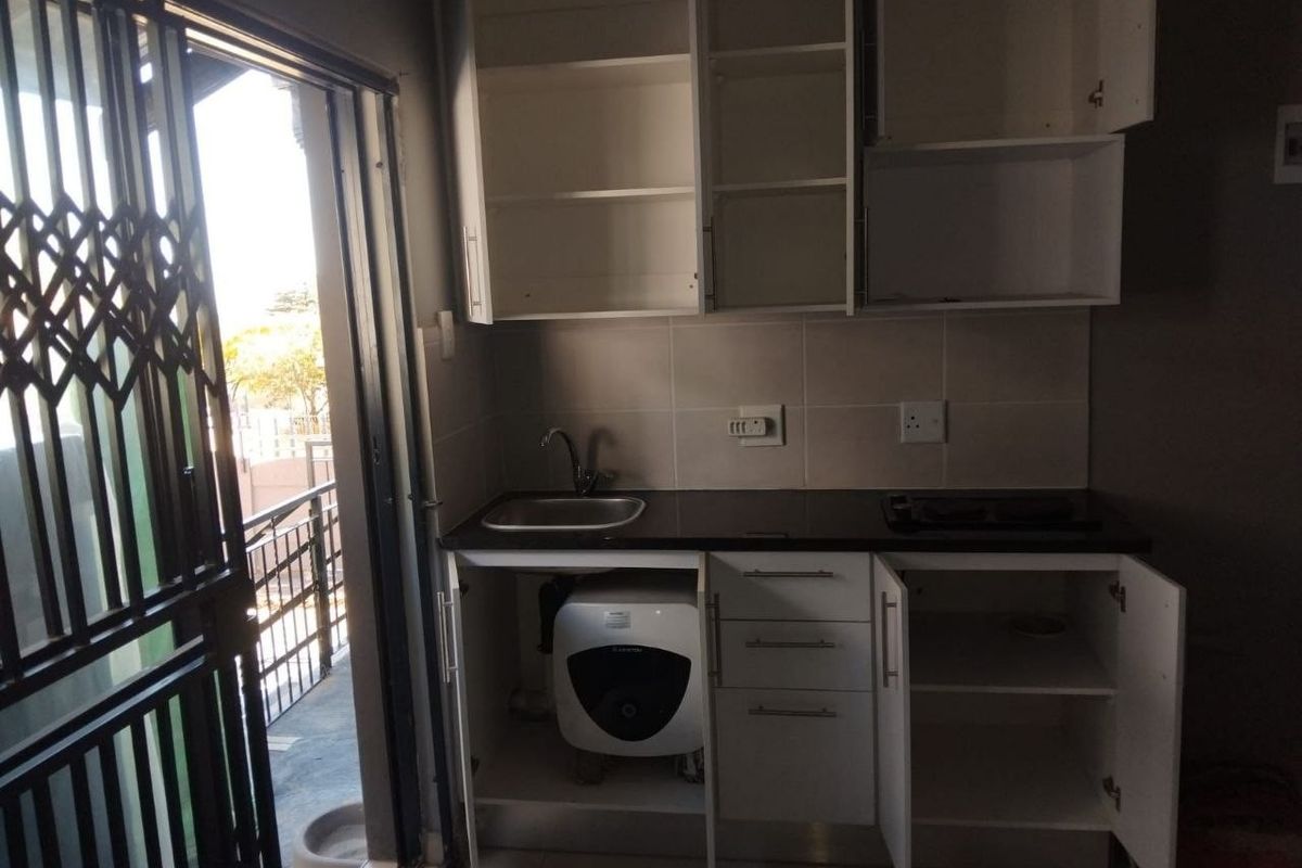 A compact kitchen area featuring built-in cabinetry, a sink, and a tiled backsplash, with direct access to an outdoor balcony through a security gate.