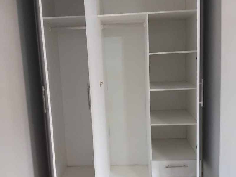 A clean, modern bedroom featuring a white built-in wardrobe with integrated shelving and drawers, complemented by neutral tiled flooring.