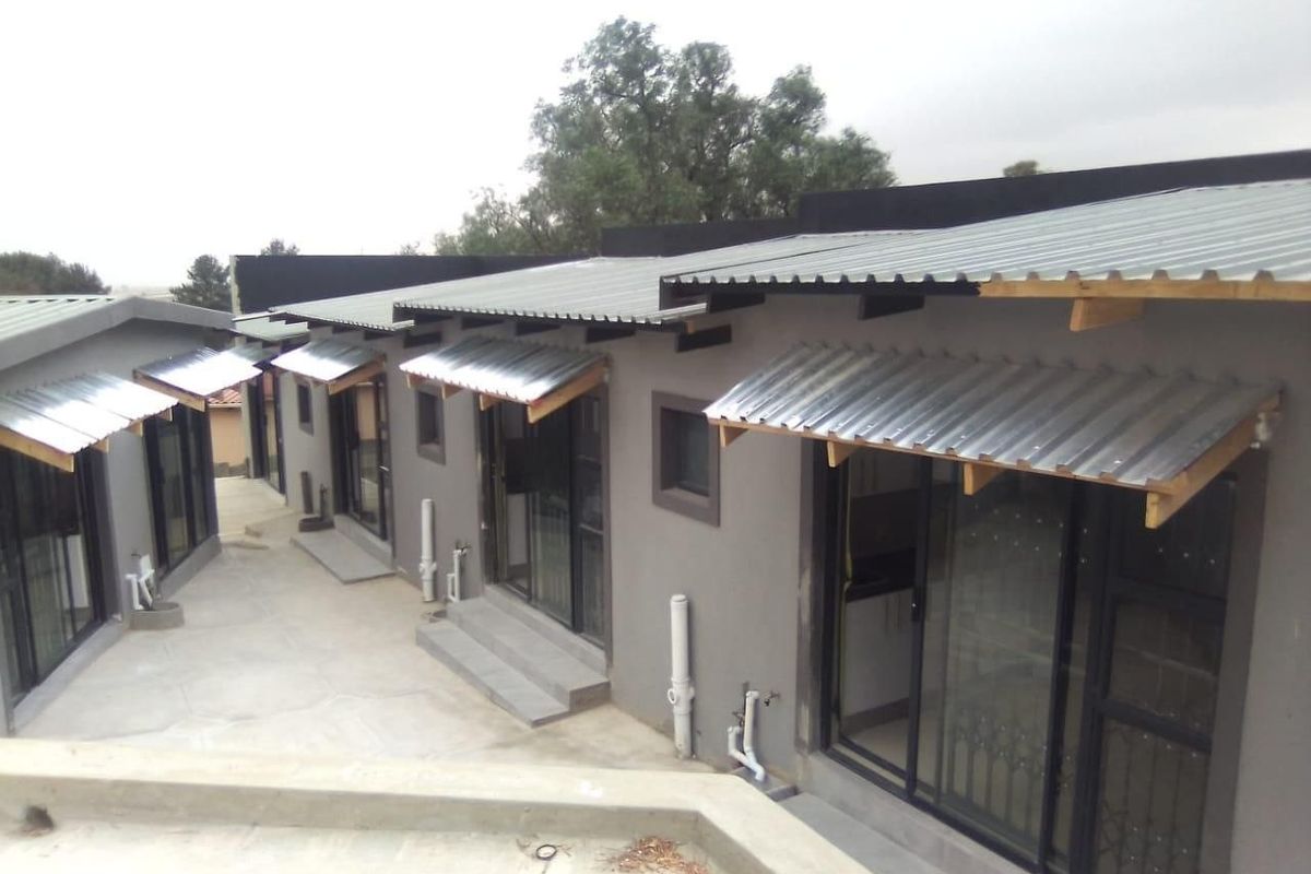Rear view of a modern residential building featuring multiple sliding glass door entrances, corrugated metal awnings, and a concrete patio area.