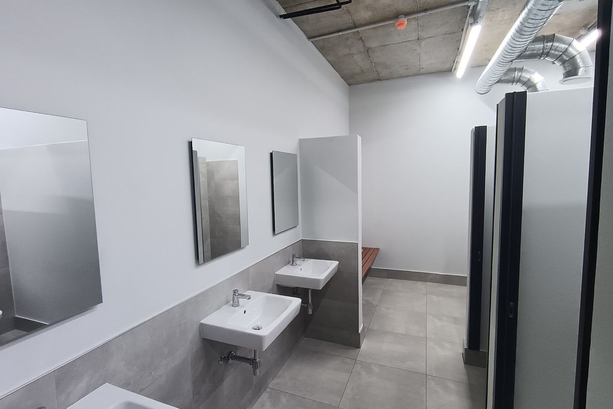 Modern industrial-style communal bathroom featuring wall-mounted basins, tiled finishes, partitioned cubicles, and exposed ceiling infrastructure with integrated lighting.
