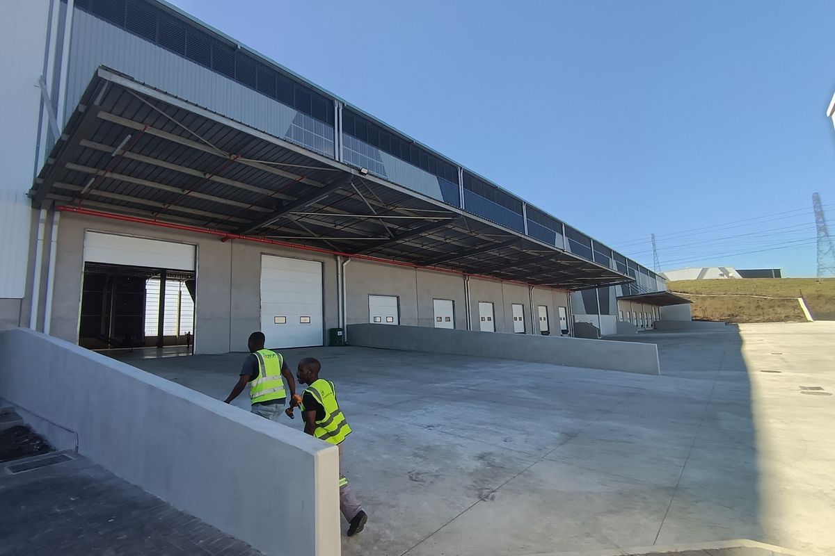 Industrial warehouse exterior featuring multiple loading docks with roller shutter doors, a large concrete yard, and an extensive overhead metal canopy.