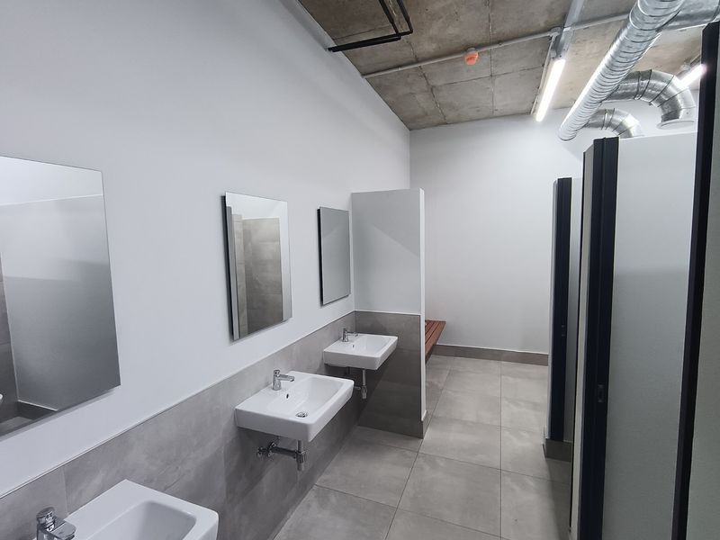 Modern industrial-style communal bathroom featuring wall-mounted basins, tiled finishes, partitioned cubicles, and exposed ceiling infrastructure with integrated lighting.