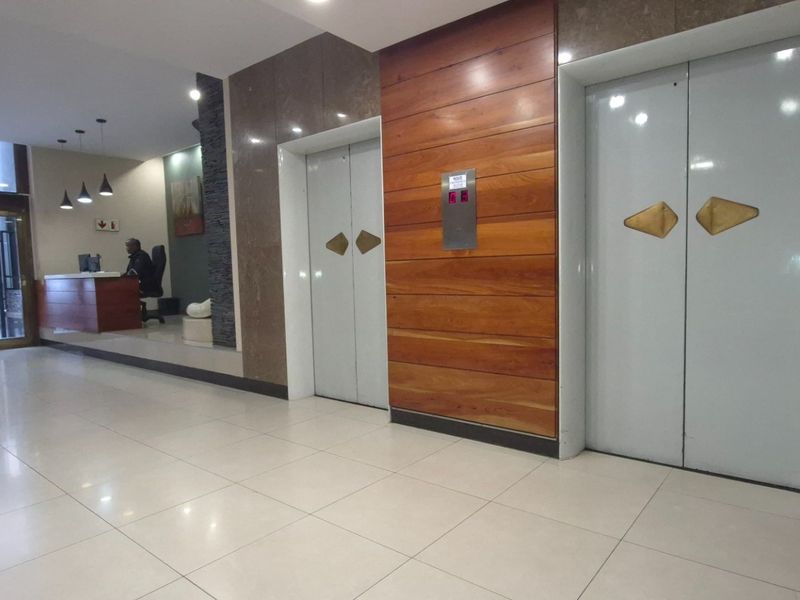 A professional commercial building lobby featuring dual elevators, wood-paneled accent walls, marble cladding, and a dedicated reception area with glass entrance doors.