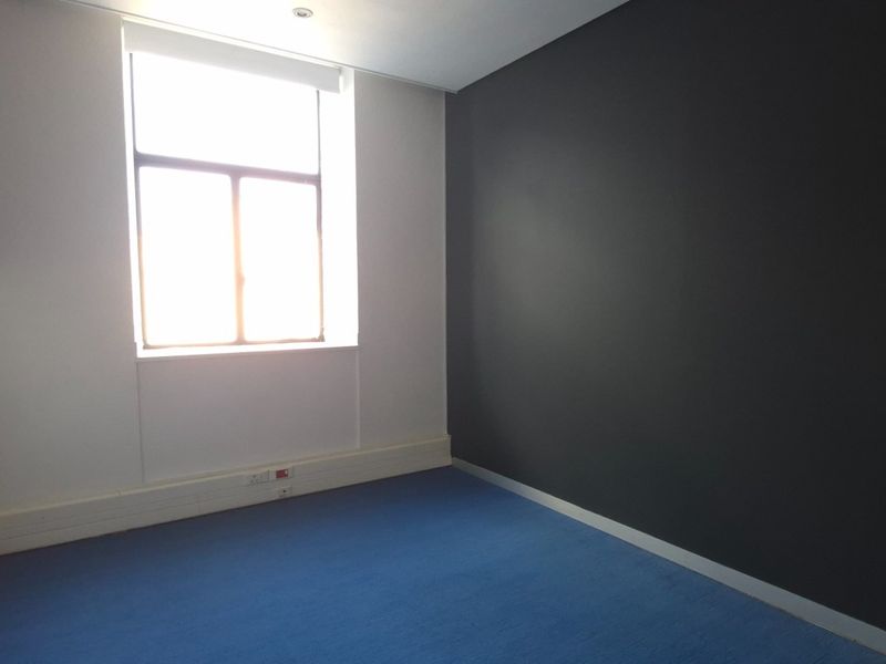 A vacant office space featuring blue carpet flooring, a large window for natural light, and a contrasting dark accent wall.