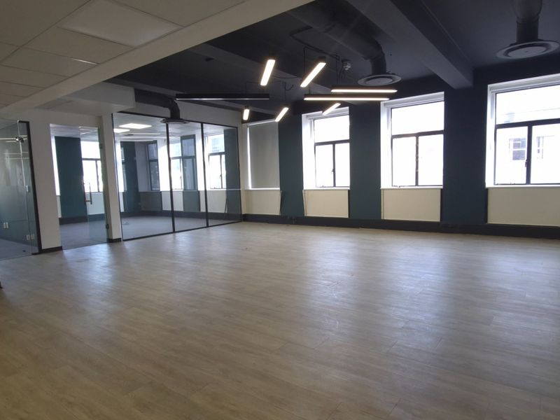 A spacious, modern open-plan office area featuring glass partitions, contemporary pendant lighting, exposed ceiling ductwork, and durable laminate flooring.