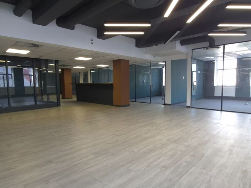 Modern open-plan commercial office space featuring glass-partitioned meeting rooms, laminate flooring, and contemporary exposed ceiling with integrated linear lighting.