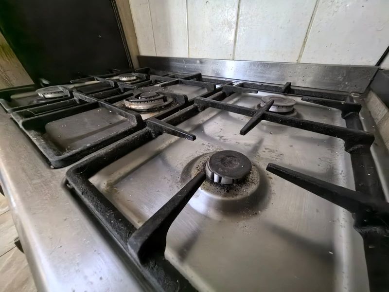 Close-up view of a stainless steel gas cooktop with cast iron trivets, showing significant signs of wear and heavy residue buildup.