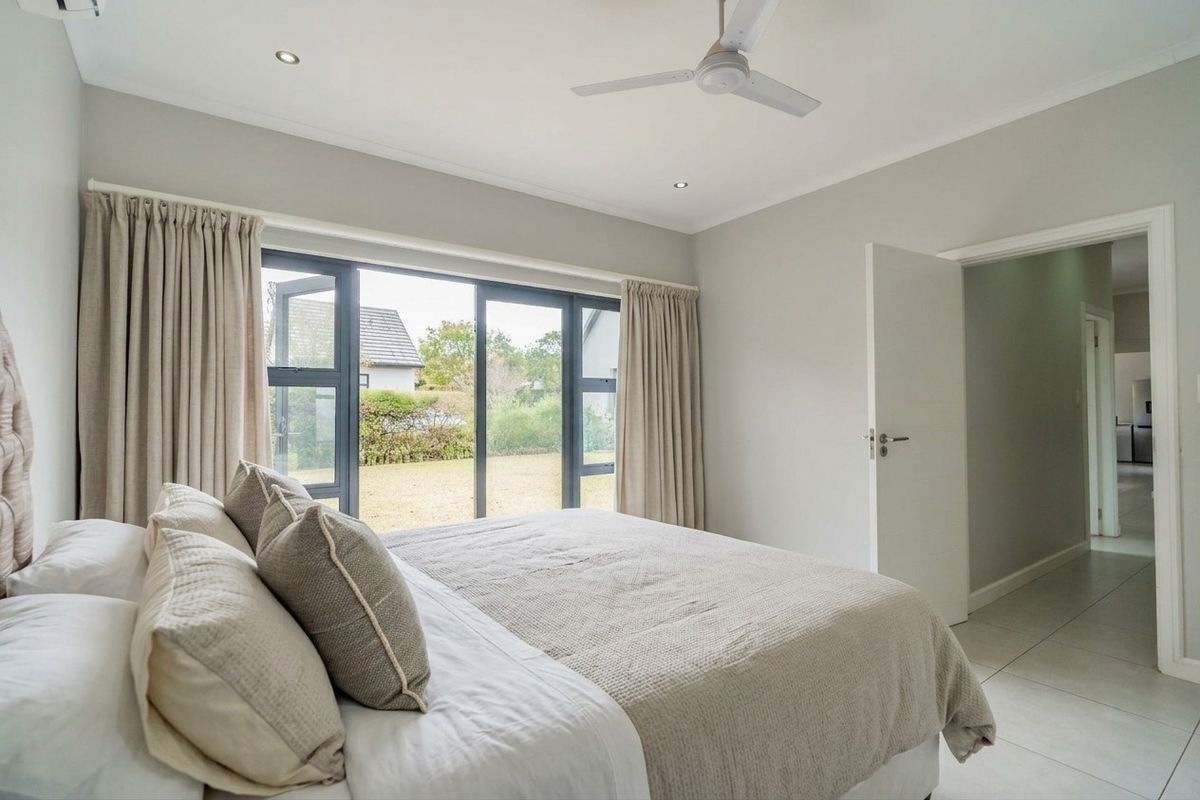 A well-maintained bedroom featuring neutral tones, tiled flooring, a ceiling fan, and large aluminium-framed windows providing natural light and garden views.