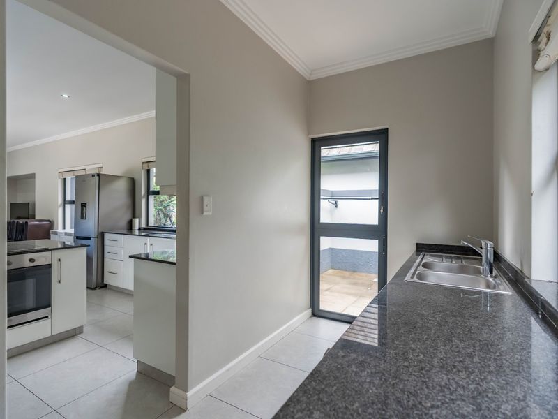 A clean and functional scullery area featuring granite countertops, a double sink, tiled flooring, and direct access to an outdoor space.