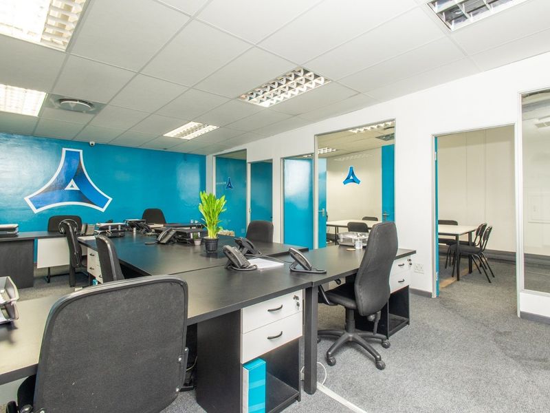 A functional open-plan office space featuring suspended ceilings, recessed lighting, carpeted flooring, and glass partition doors leading to adjacent work areas.