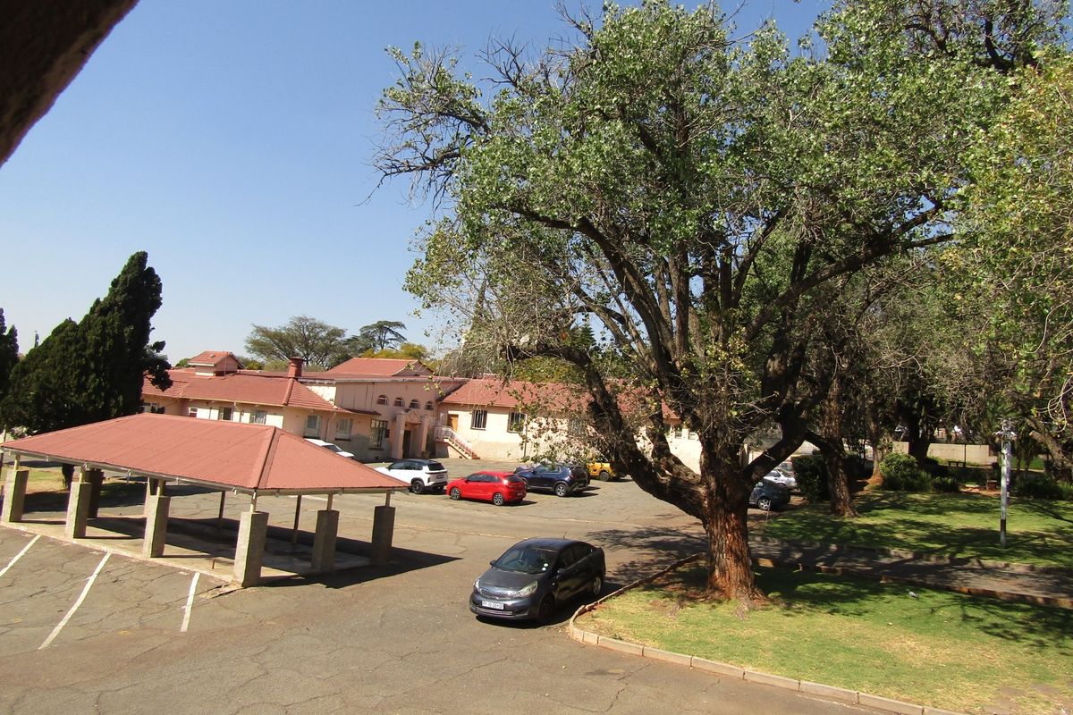 An elevated view of the apartment complex grounds, featuring a covered parking area, paved driveway, and established garden space with mature trees.