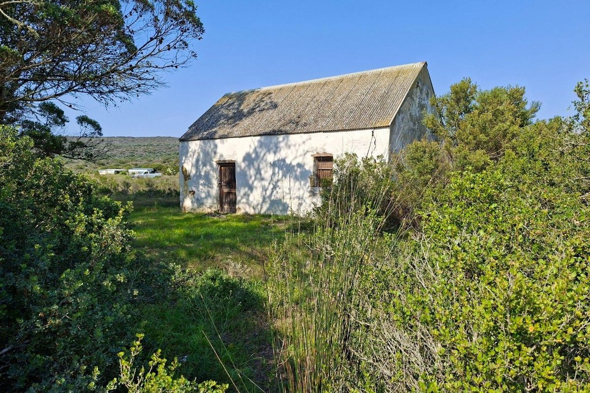 A rustic, detached farm outbuilding featuring weathered masonry walls and a corrugated roof, situated in a natural, overgrown rural landscape.