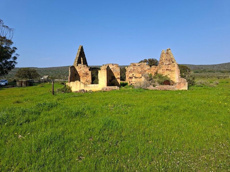 A derelict stone farmhouse ruin situated on a large open field, showcasing original masonry walls and a collapsed roof structure.