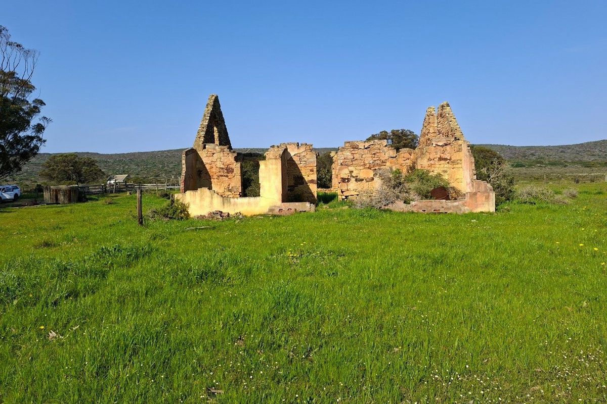 A derelict stone farmhouse ruin situated on a large open field, showcasing original masonry walls and a collapsed roof structure.