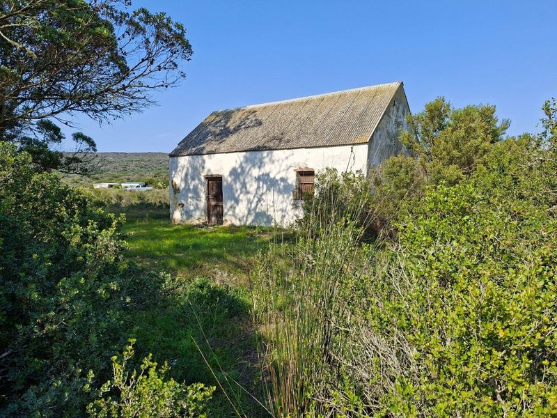 A rustic, detached farm outbuilding featuring weathered masonry walls and a corrugated roof, situated in a natural, overgrown rural landscape.