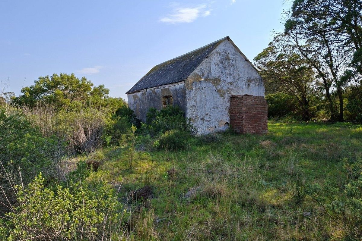 A weathered, detached farm outbuilding featuring a pitched roof and brick chimney, situated on a rural plot with significant surrounding vegetation.