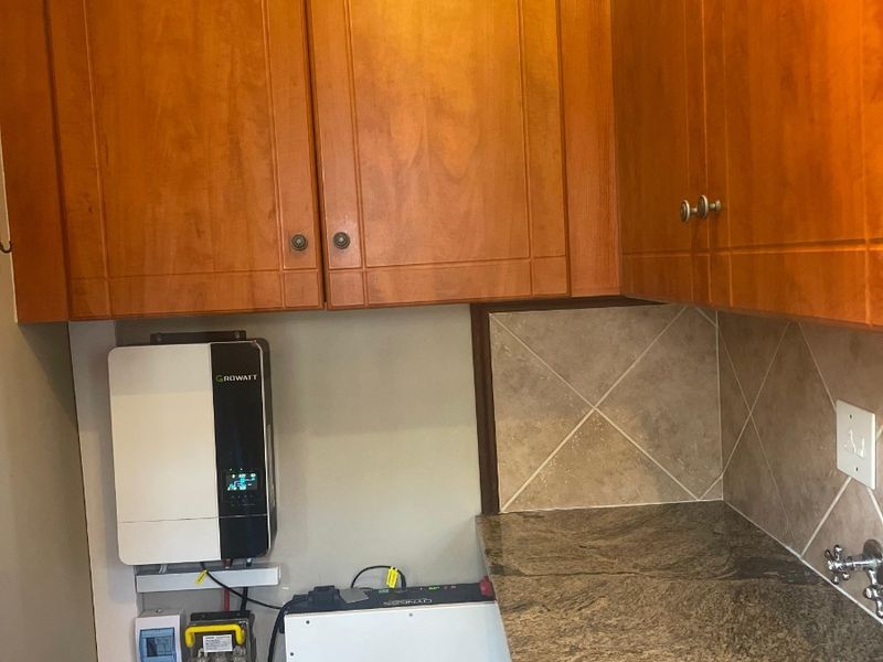 A utility area featuring granite countertops, tiled backsplash, wooden cabinetry, and a wall-mounted solar inverter with battery storage system.
