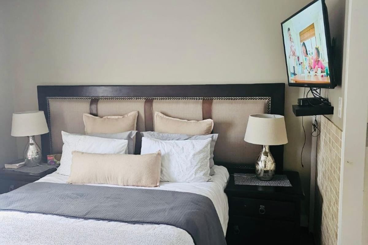A comfortable bedroom featuring a wall-mounted air conditioning unit and a television mount, with neutral wall tones and tiled accent detailing.