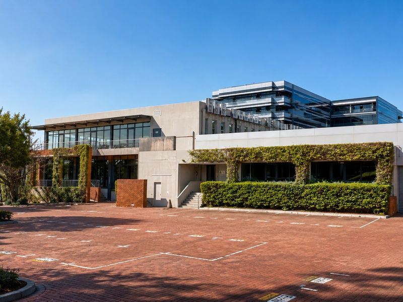 Exterior view of a modern office building featuring a large paved parking area, roller shutter door, and landscaped climbing greenery.